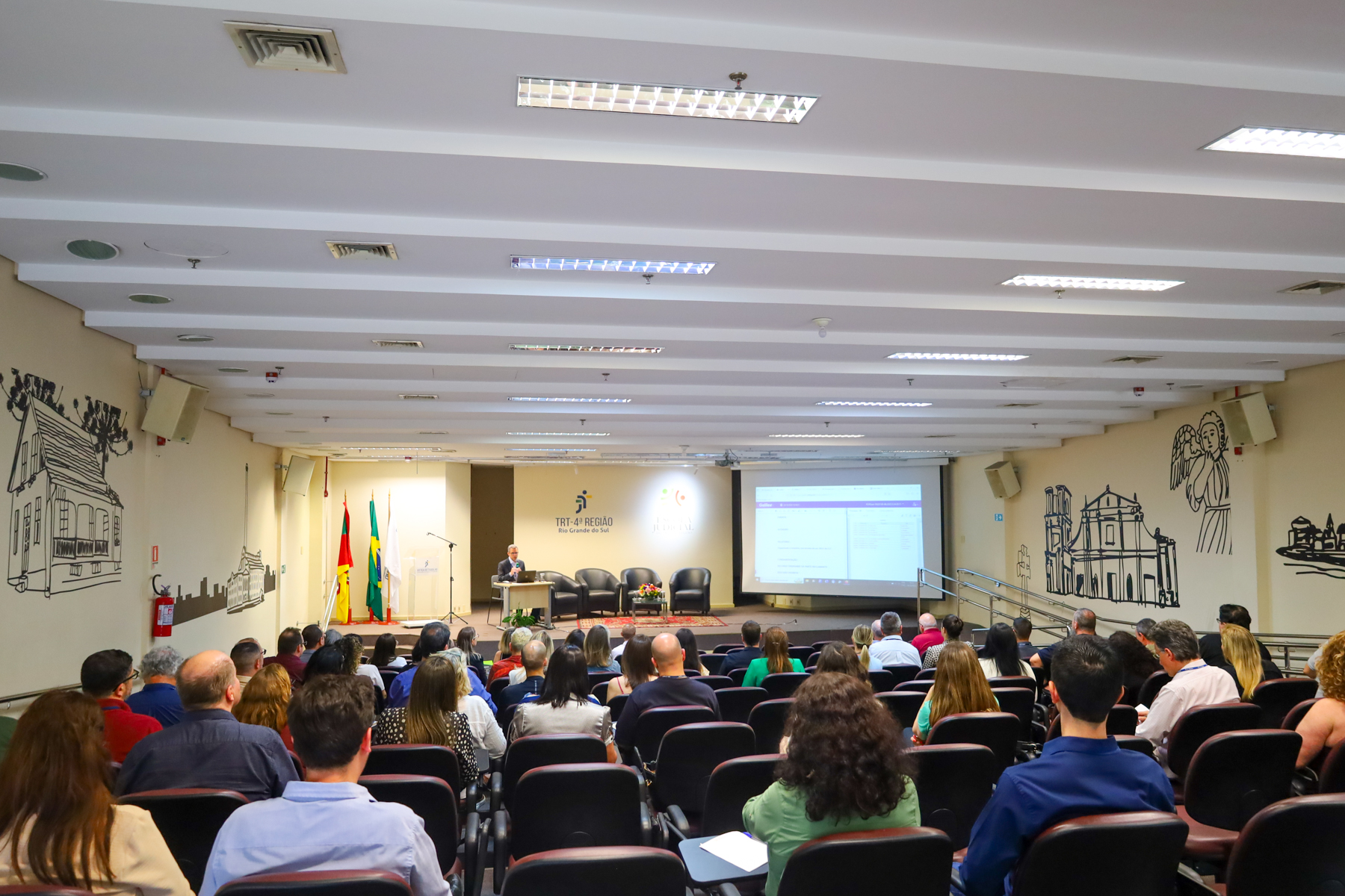 Frederico Cardoso, falando no palco. Ao lado dele, o telão com a apresentação. Público aparece em primeiro plano, de costas.