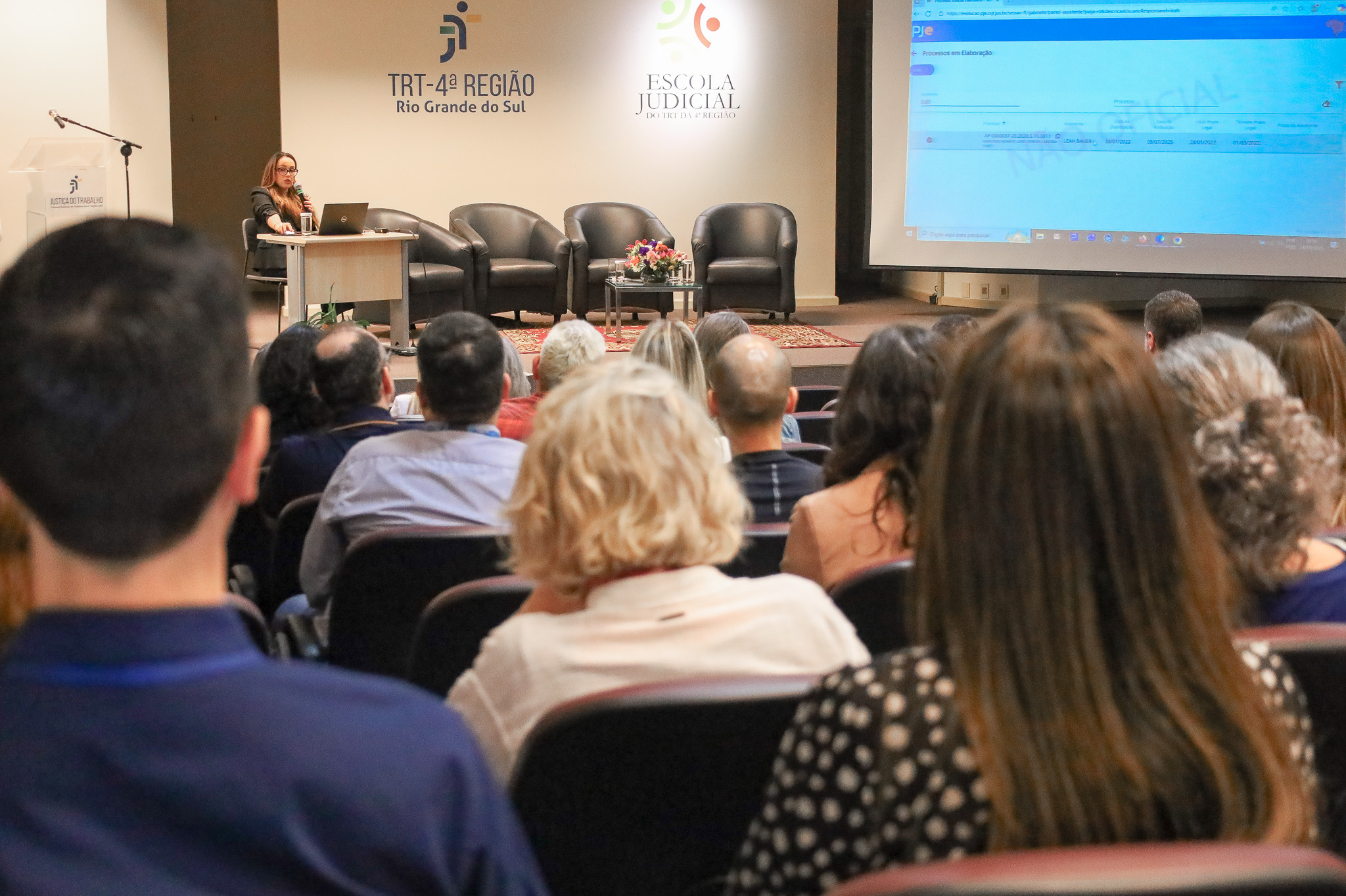 No palco, Mileice Correa falando. Público aparece em primeiro plano, de costas.