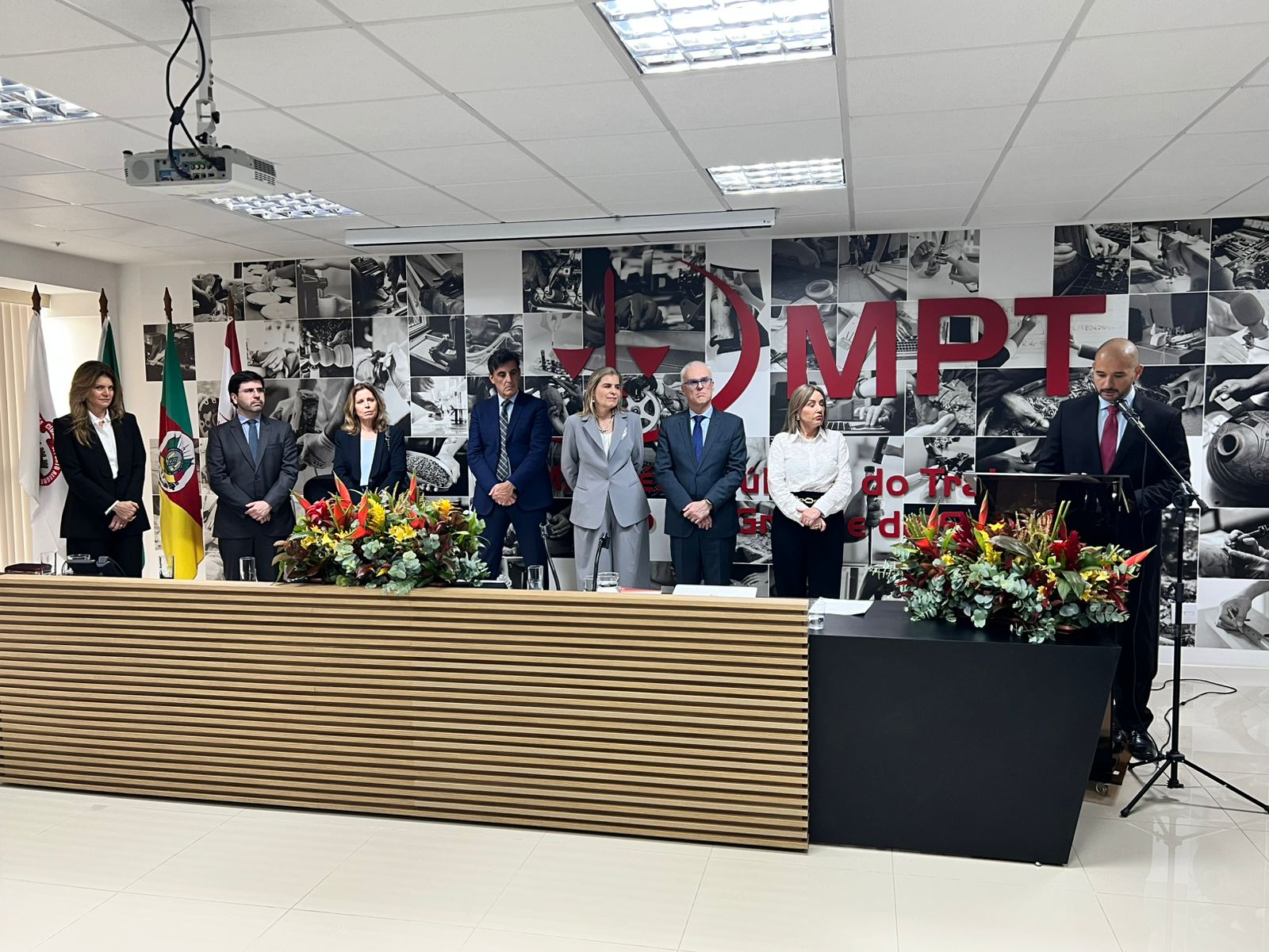 Antônio Pereira está de pé, no canto direito da foto, falando ao microfone no púlpito. Na esquerda, sete pessoas da mesa oficial aparecem também de pé acompanhando sua fala. Ao fundo aparece o logo do MPT