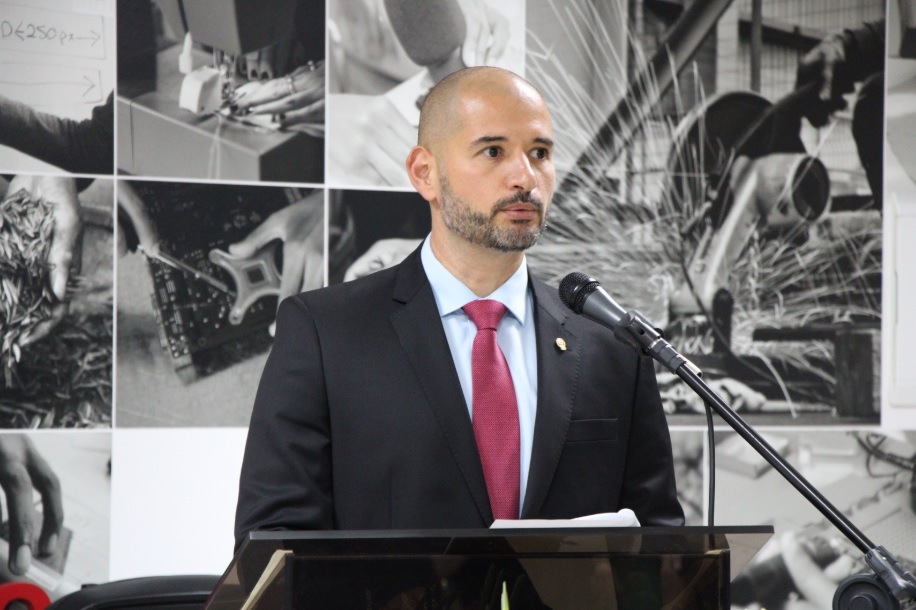 Antônio Pereira é branco, calvo e usa barba curta. Veste terno preto e gravata vermelha com camisa branca. Está falando ao microfone e olhando para o lado direito da imagem. Ao fundo aparece um mosaico de imagens representando diferentes formas de trabalho. 