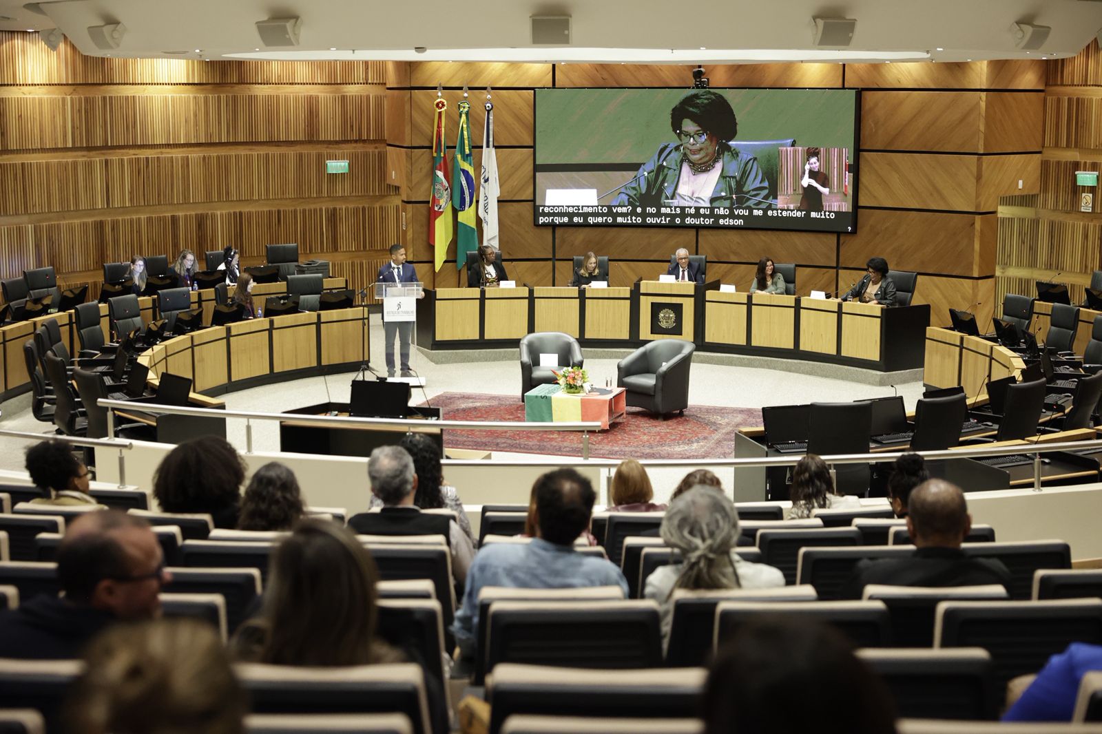 Foto em ângulo aberto, mostrando público em primeiro plano, de costas, e a mesa oficial e o telão, ao fundo.