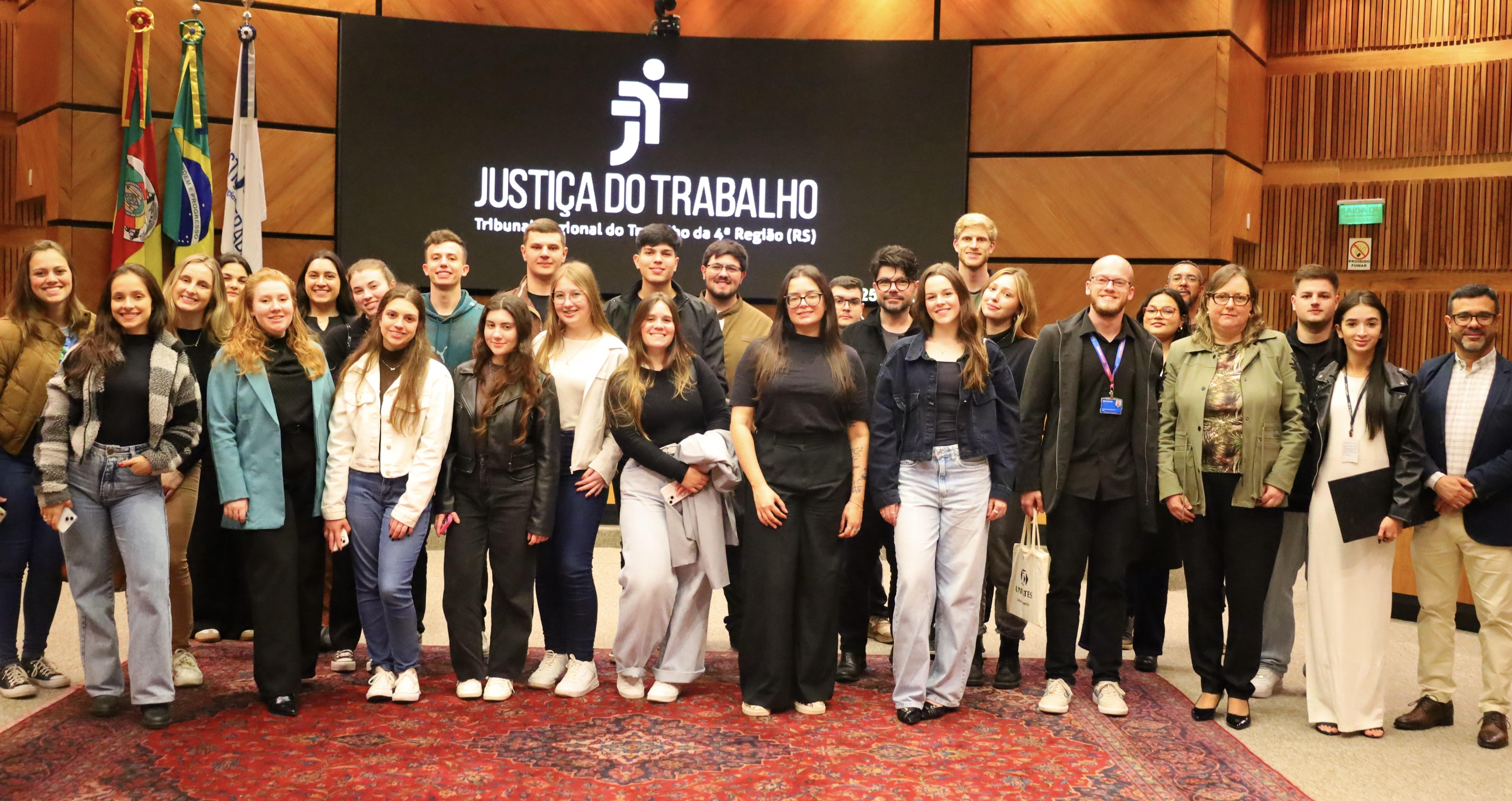 Fotografia dos estudantes no plenário em frente ao painel escrito “Justiça do Trabalho – Tribunal Regional do Trabalho da 4ª Região (RS)".