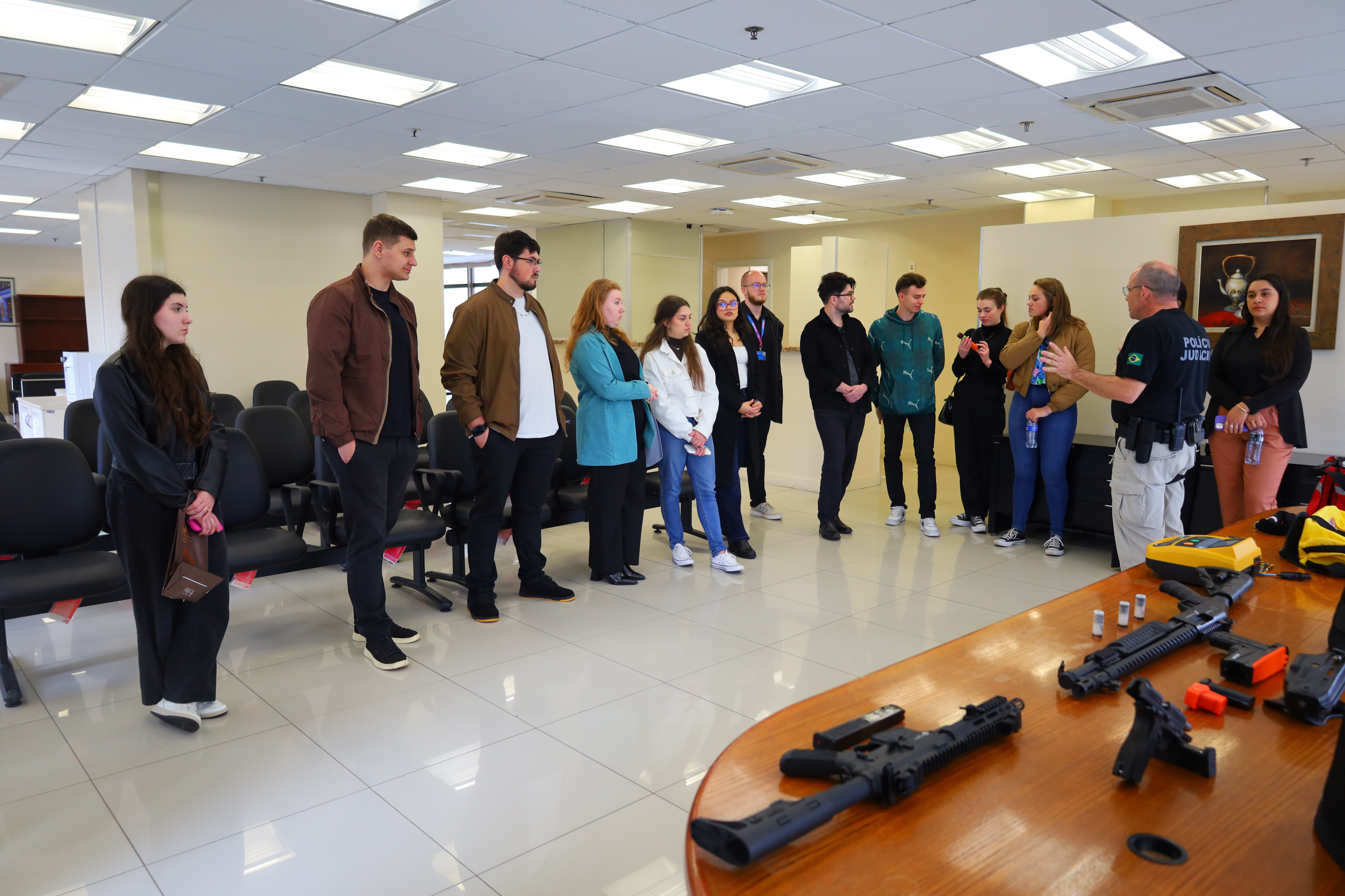 Na sala de convivência, 12 estudantes estão de pé escutando a explicação de Marcelo, há equipamentos de porte leve sobre a mesa.