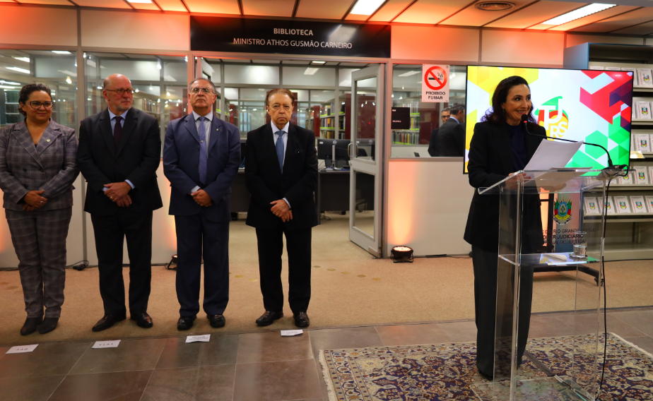 Representantes das instituições aparecem lado a lado. Mais à frente, a mestre de cerimônias fala ao microfone. Ao fundo, vê-se a fachada de entrada da biblioteca.