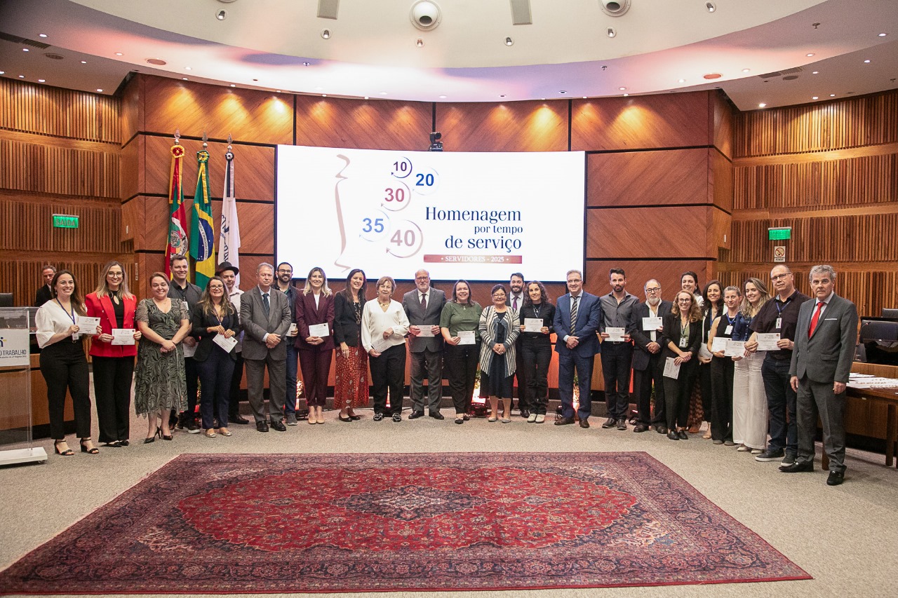 Foto posada de várias pessoas sorrindo e olhando para a câmera com os certificados de tempo de serviço. Eles estão na nave do Plenário e atrás deles aparece o telão com o logo do evento.