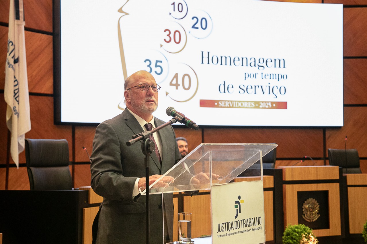 Martins Costa é branco, calvo e grisalho. Usa barba curta. Veste terno e gravata e fala ao microfone do púlpito de pé. atrás dele aparece o telão com a  arte do evento.