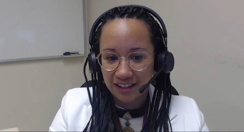 Roberta é uma mulher negra, jovem. Ela tem cabelos longos, trançados. Veste casaco e blusa brancos, óculos de grau com armação transparente e headphone.