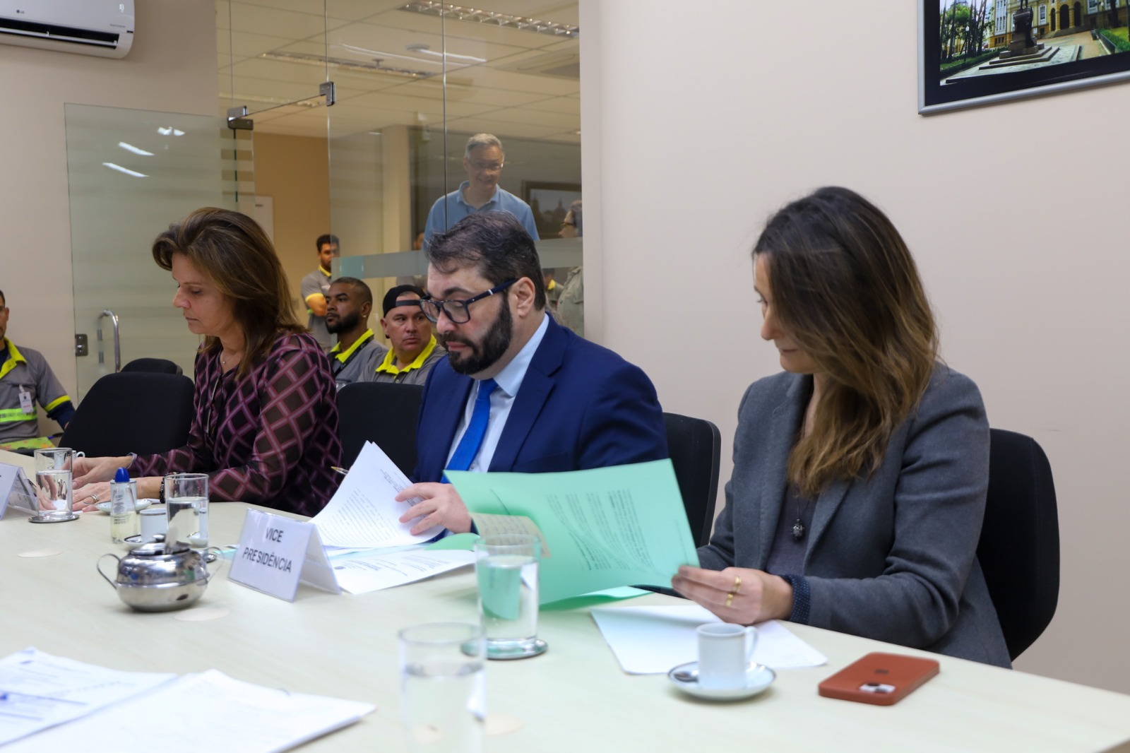 A foto mostra três pessoas em primeiro plano sentadas da mesa.