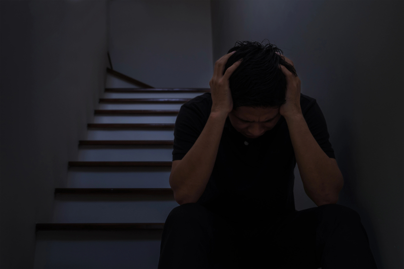Homem sentado em escada segura a cabeça, em sinal de tristeza/perturbação. Cena é escura