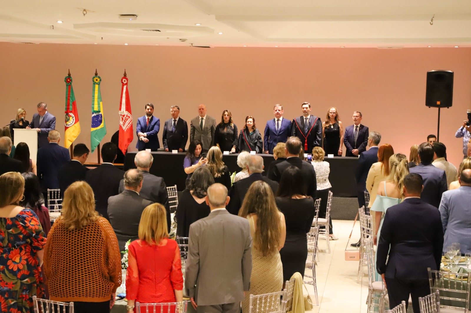 A foto mostra pessoas de pé num salão, numa cerimônia.