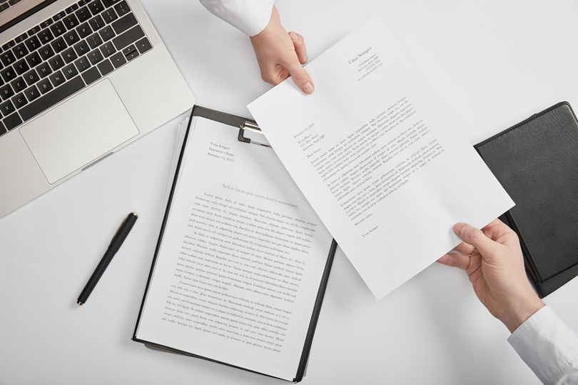 A foto ilustrativa mostra uma cena de escritório, com um fundo branco e objetos organizados sobre uma mesa de trabalho. No lado esquerdo há um notebook prateado parcialmente visível, com o teclado exposto. Abaixo dele, vê-se uma caneta preta apoiada na mesa. No centro, há uma prancheta com um documento preso, repleto de texto.