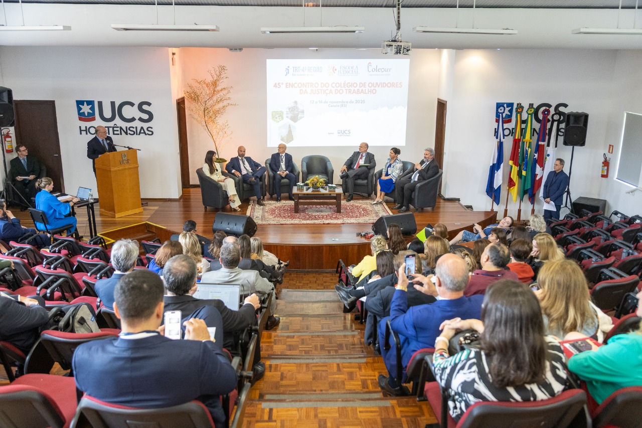 Foto geral do evento, com público em primeiro plano de costas e os palestrantes no palco ao fundo.