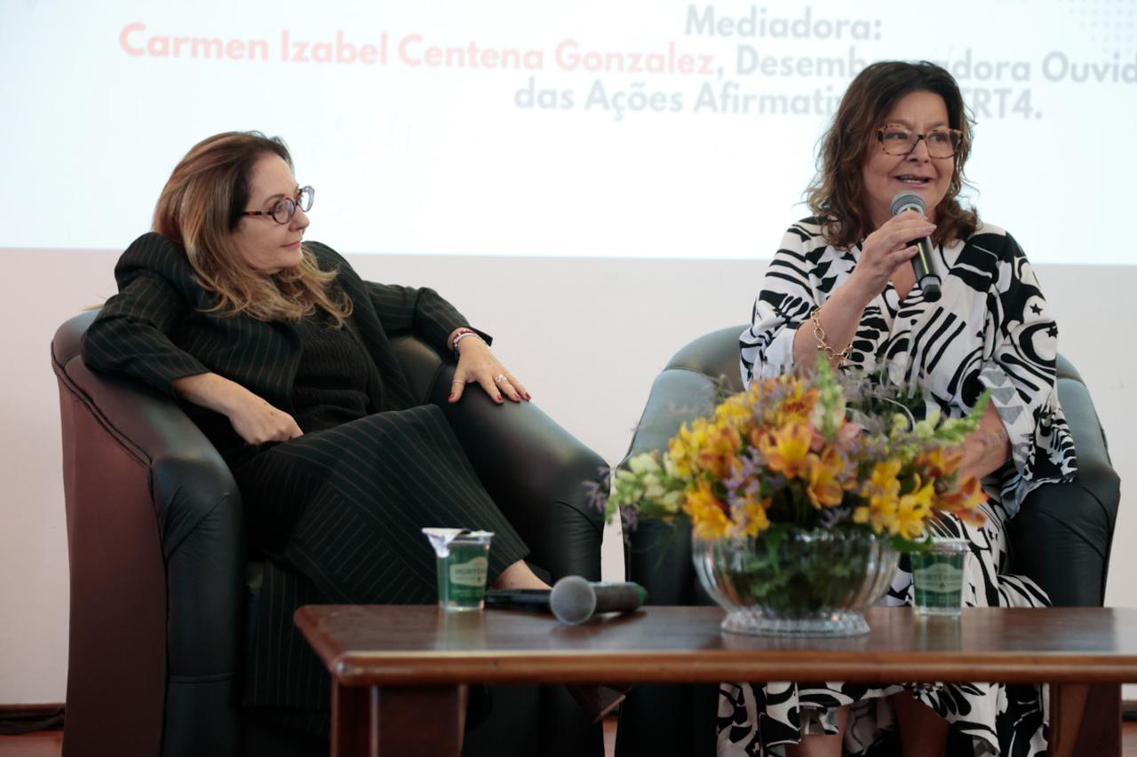 Liana e Carmen estão sentadas em poltronas no palco. Carmen fala ao microfone.