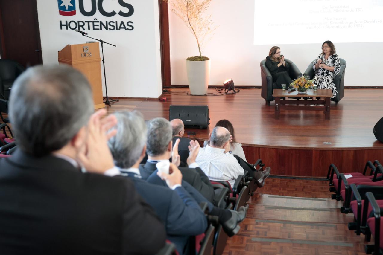 Público está de costas aplaudindo em primeiro plano, Liana e Carmen aparecem no palco ao fundo.