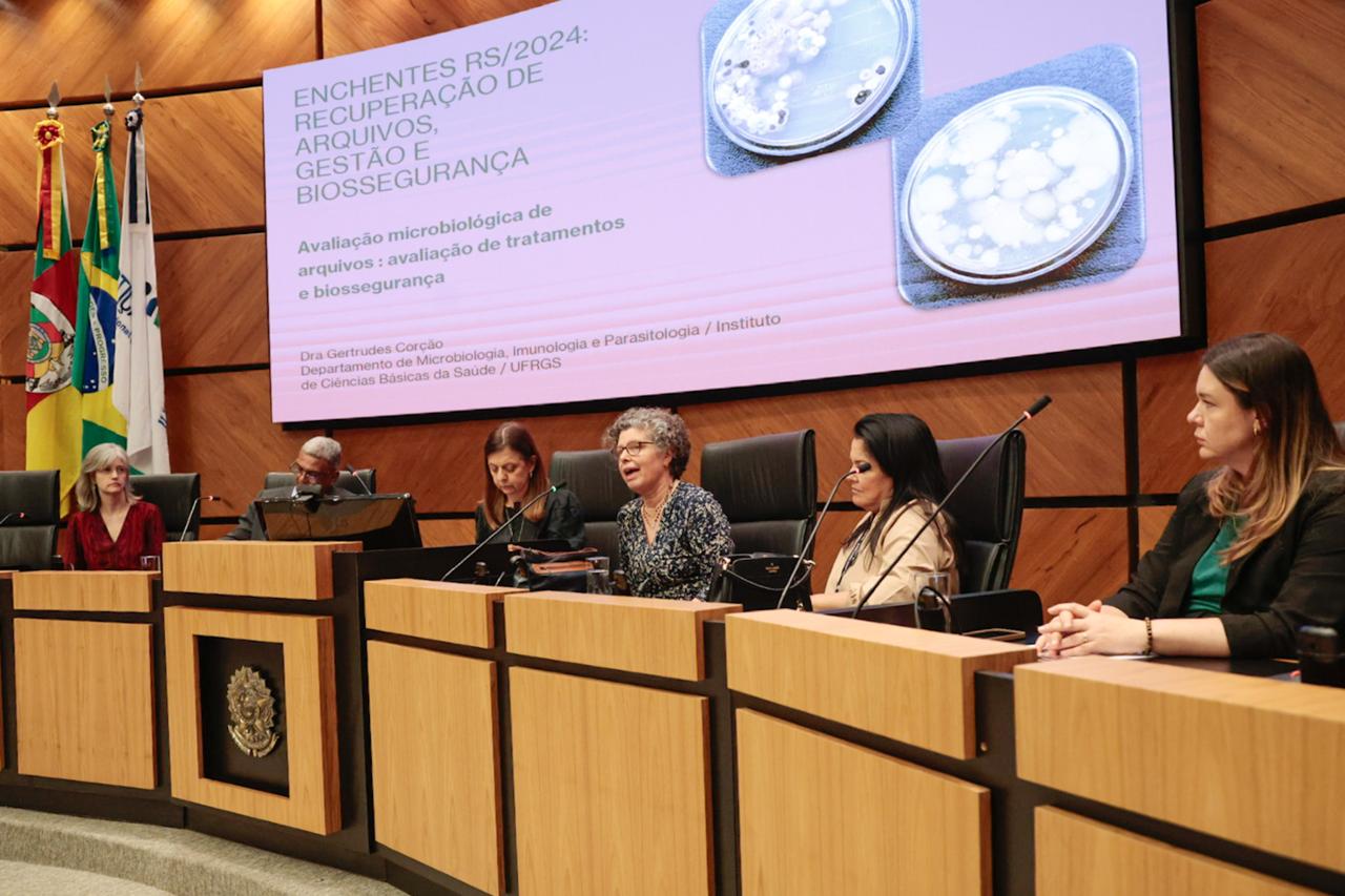 foto da mesa oficial, uma mulher fala ao microfone ao lado de cinco pessoas.