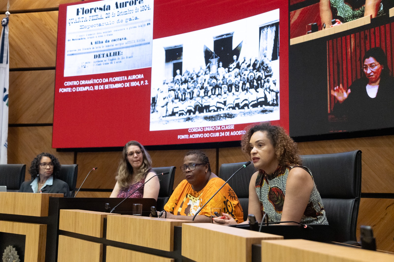 Fernanda é negra, com cabelos crespos até os ombros. Usa camisa com etampas em preto, branco e vermelho. Fala ao microfone em primeiro plano. A sua esquerda, três mulheres aparecem na bancada ouvindo sua fala.