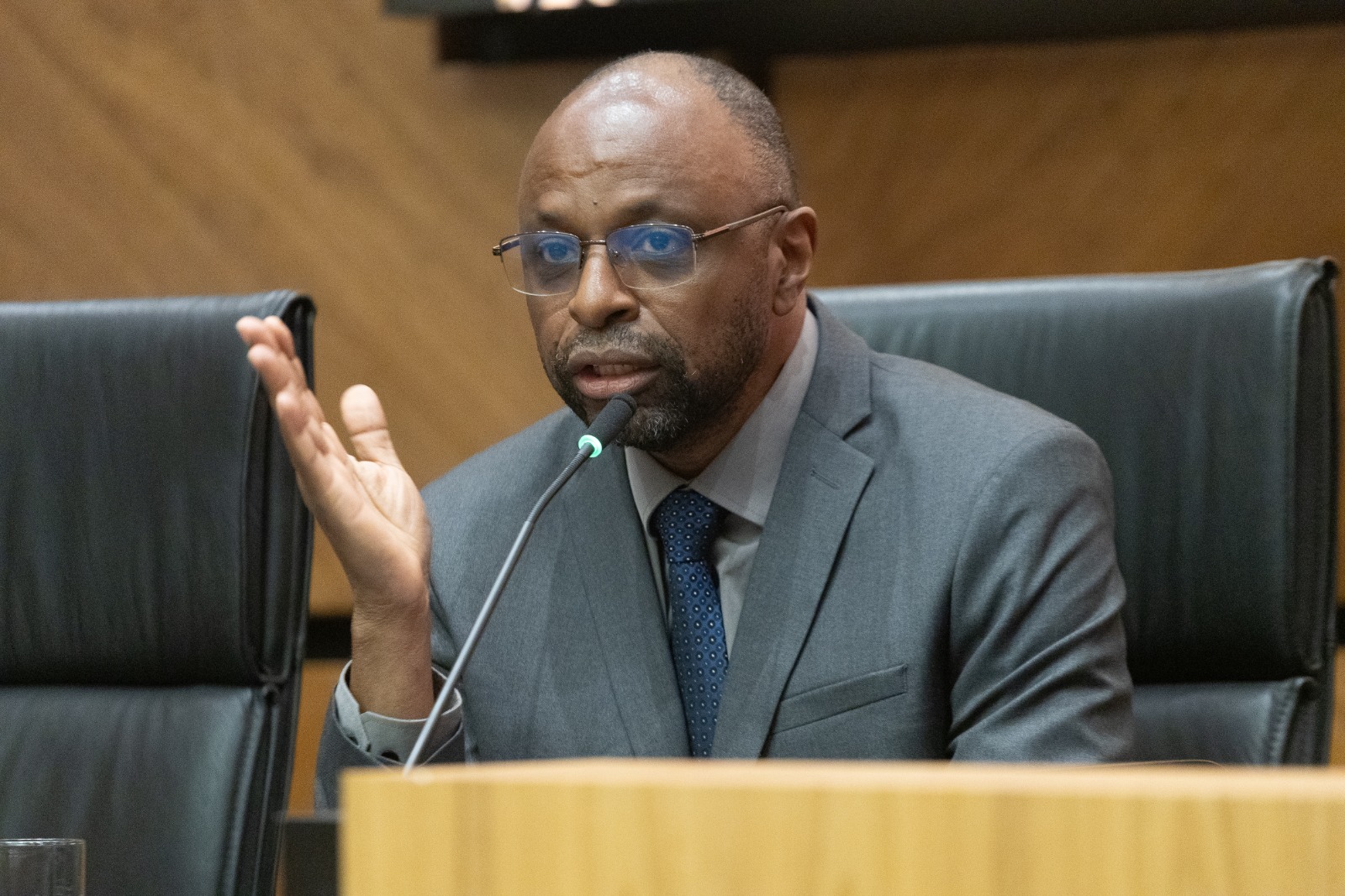 Lilian é negra, usa camisa com estampas em preto e branco, fala gesticulando e olhando para a esquerda da imagem. Usa óculos de aro grande e o cabelo preto preso em coque. A sua esquerda, duas pessoas aparecem na bancada, fora de foco e de costas, ouvindo sua fala.