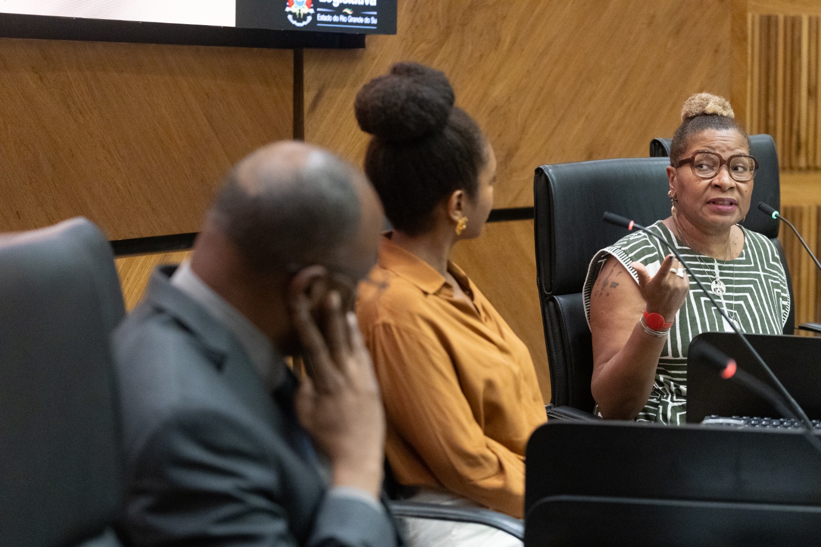 Lilian é negra, usa blusa com estampa em branco e preto, cabelo preto em coque e óculos de aro grande. Fala e gesticula olhando para a esquerda da imagem. a sua esquera, duas pessoas da bancada aparecem de costas e fora de foco ouvindo sua fala. 