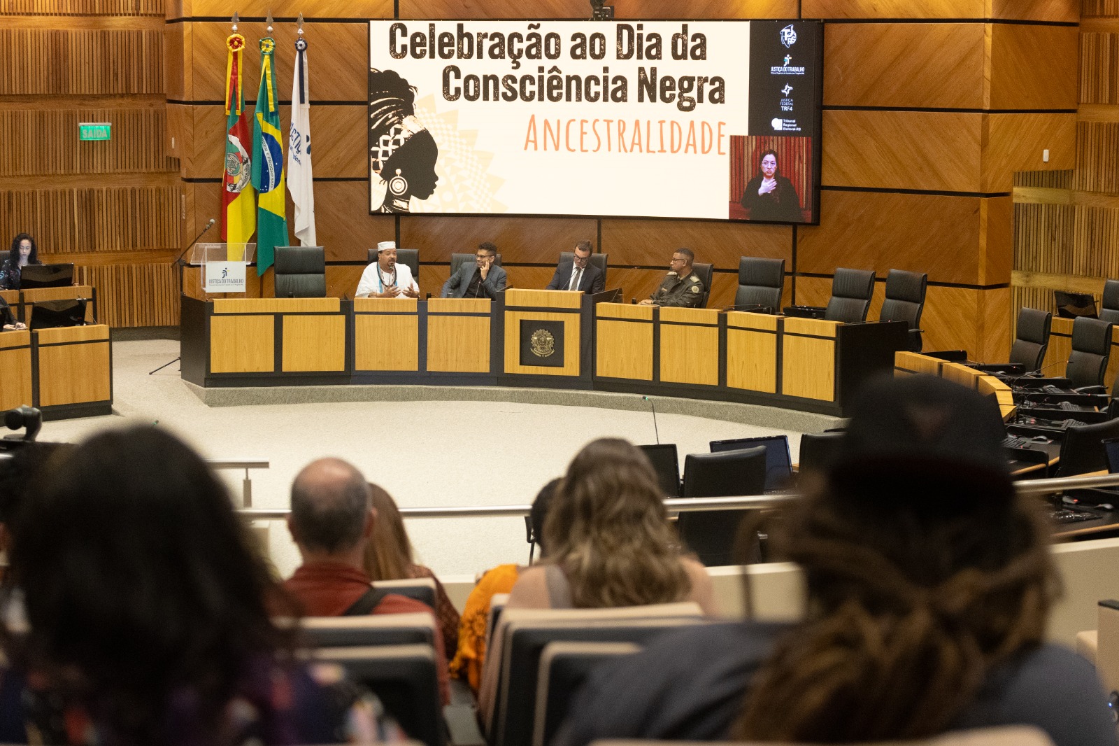 Nave do Plenário com quatro pessoas na bancada. Ao fundo aparece um telão ligado com a arte do evento. Em primeiro plano, pessoas do público assistem de costas.