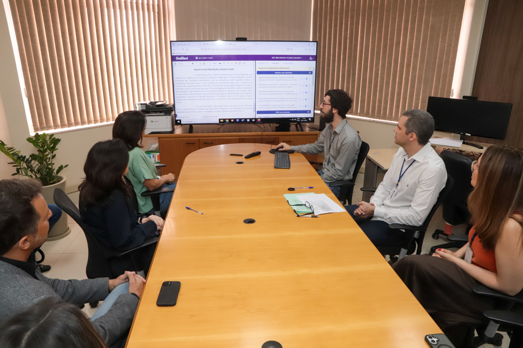 foto da reunião, mostrando participantes sentados à mesa. Ao fundo, o telão com o sistema.