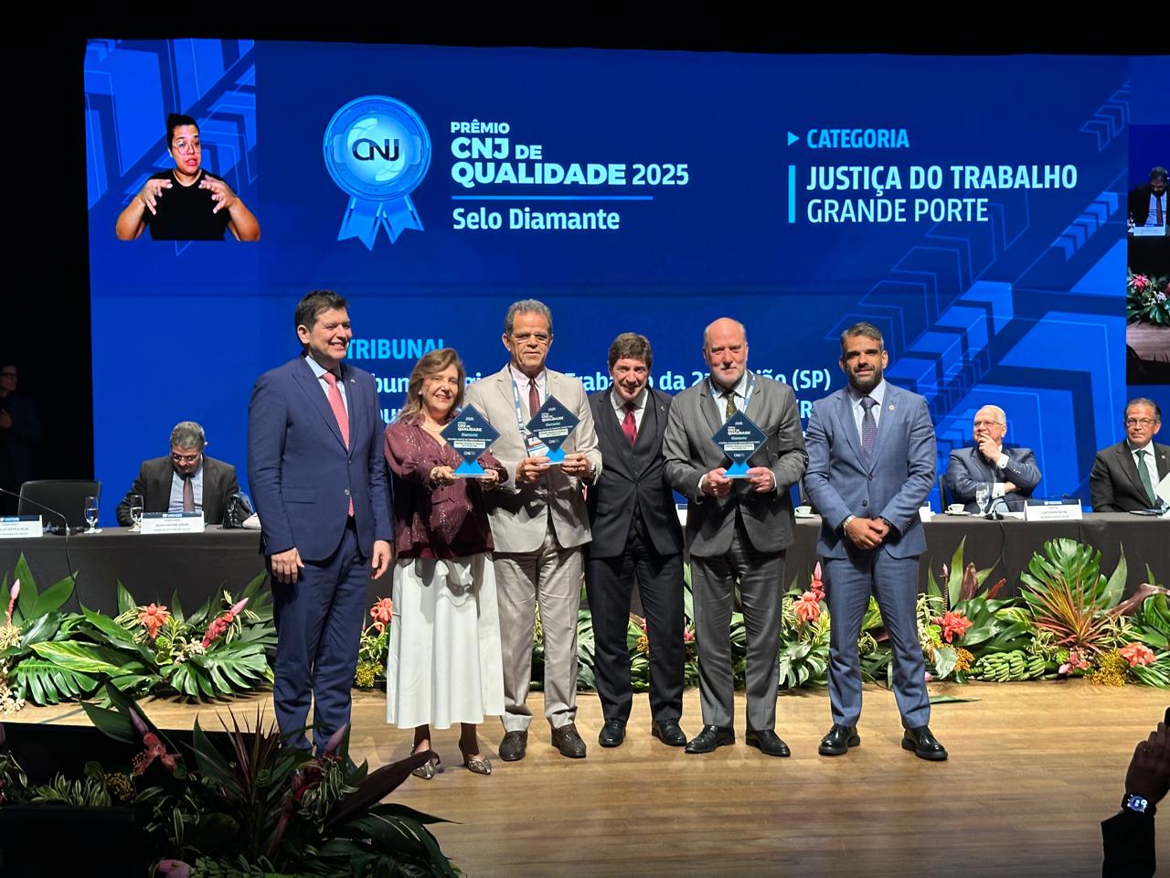 Seis pessoas aparecem no palco. Um telão ao fundo exibe a arte do evento. 
