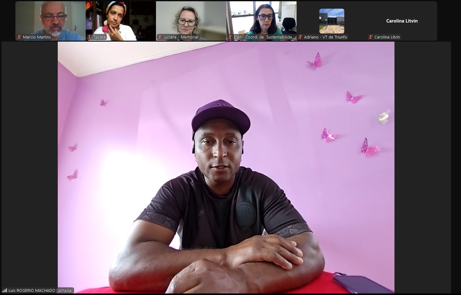 Rogério é negro, calvo, usa boné roxo, camiseta preta com detalhes em cinza. Está de braços cruzados sobre a mesa e fala para a câmera. 