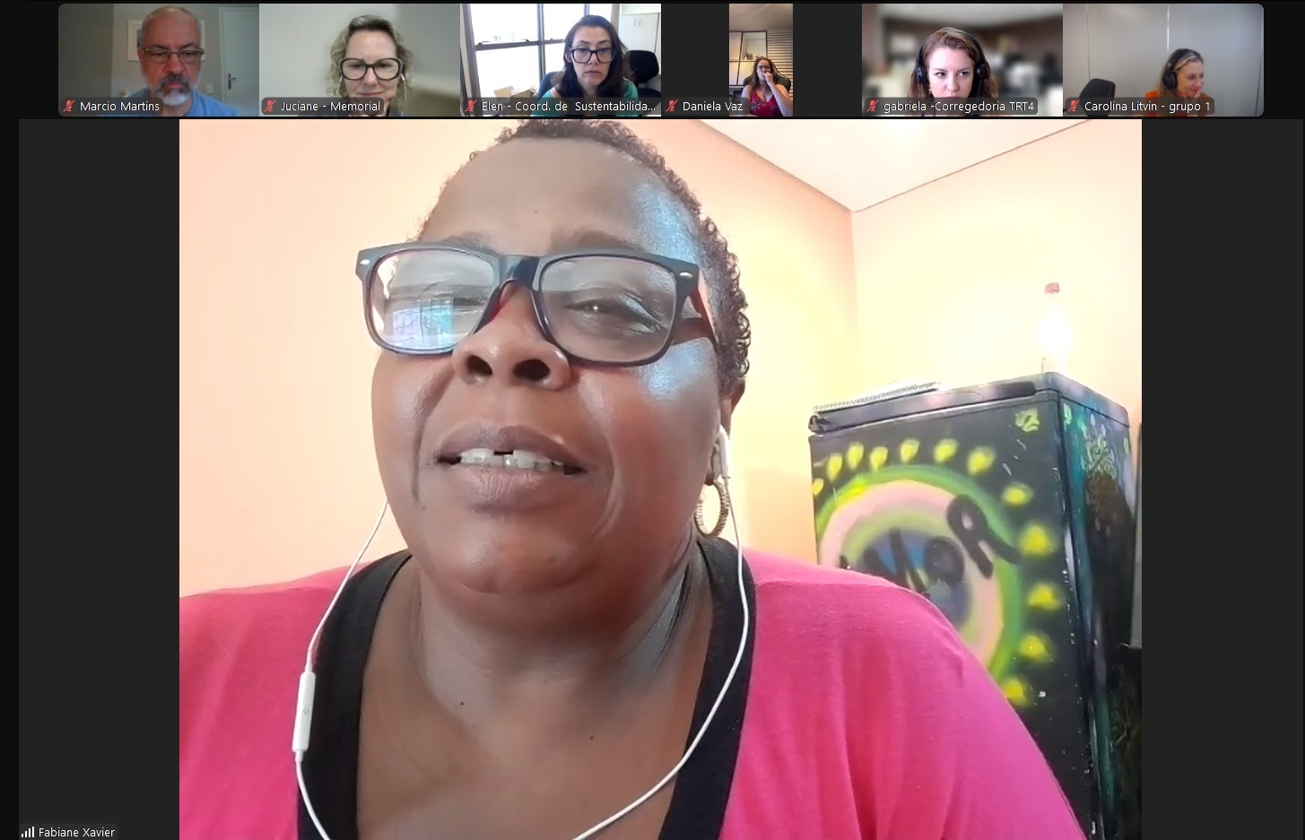 Fabiane é negra, usa cabelo curto, óculos de aro grande e vestido rosa. ao fundo, aparece um geladeira com um pintura onde se lê "amor". 