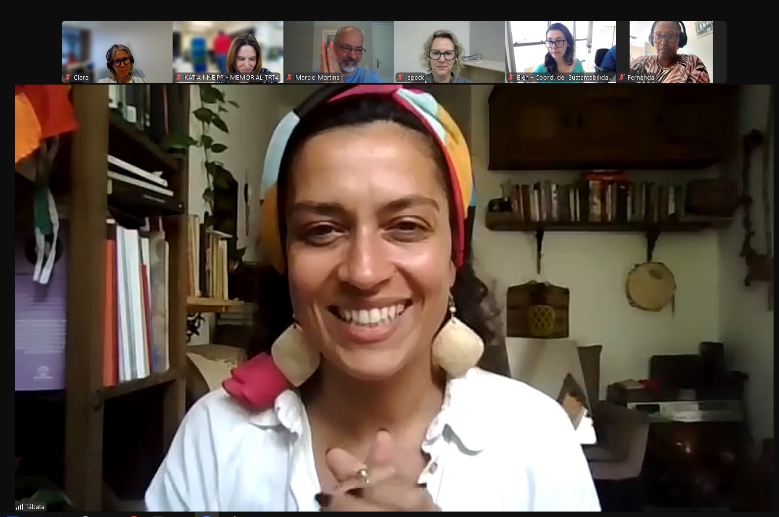 Tábata é negra, usa um lenço colorido sobre os cabelos. Está sorrindo, usa camisa branca. Ao fundo aparecem diversos objetos de decoração na parede.