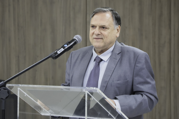 Edson é branco, de cabelos grisalhos. Fala de pé ao microfone. Usa terno cinza, camisa cinza claro e gravata xadrez