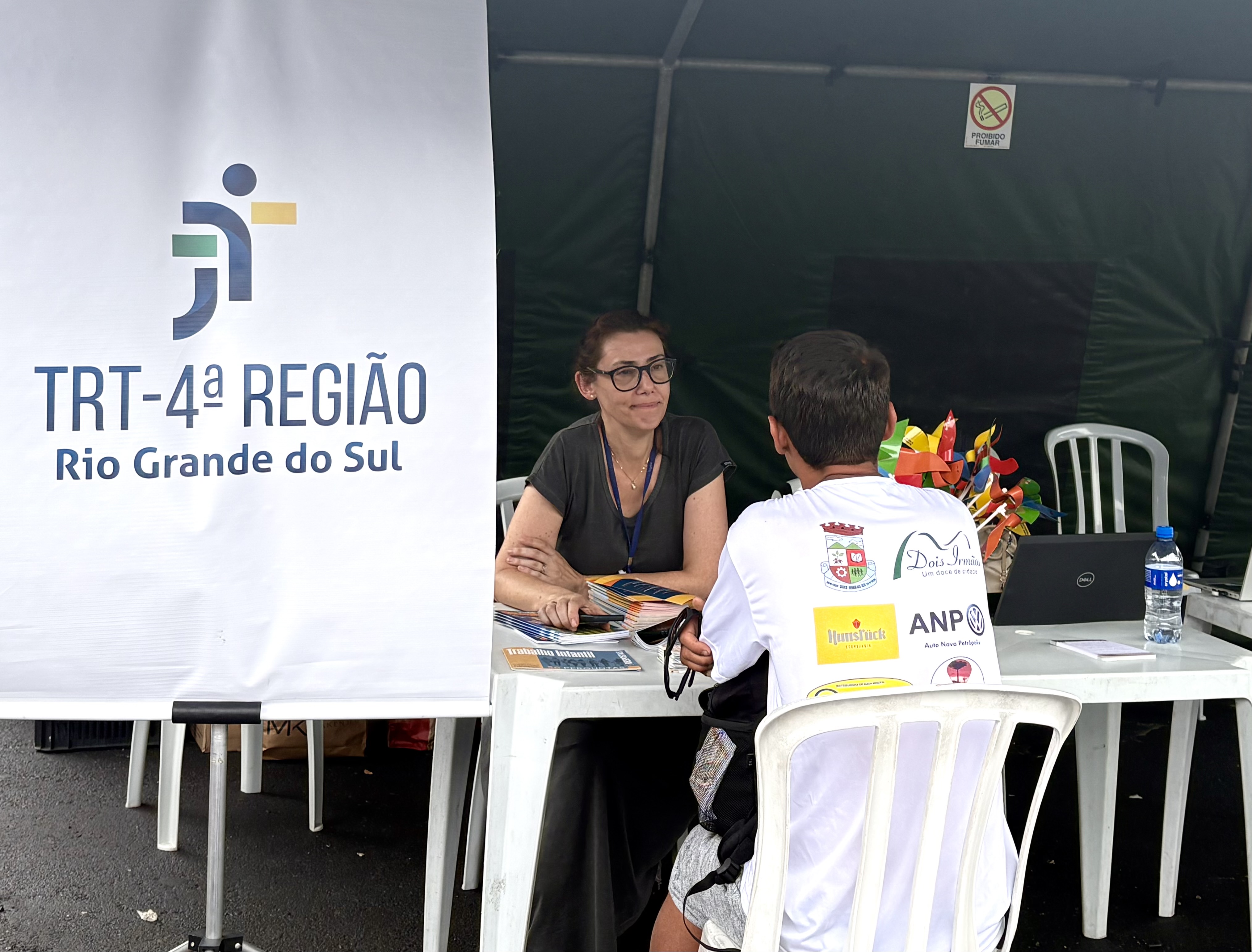 A servidora Elen do TRT-RS realiza atendimento individual em uma mesa, conversando com um cidadão sentado à sua frente. Ao lado, há um banner do TRT. Sobre a mesa, estão materiais informativos, computador e garrafa de água.
