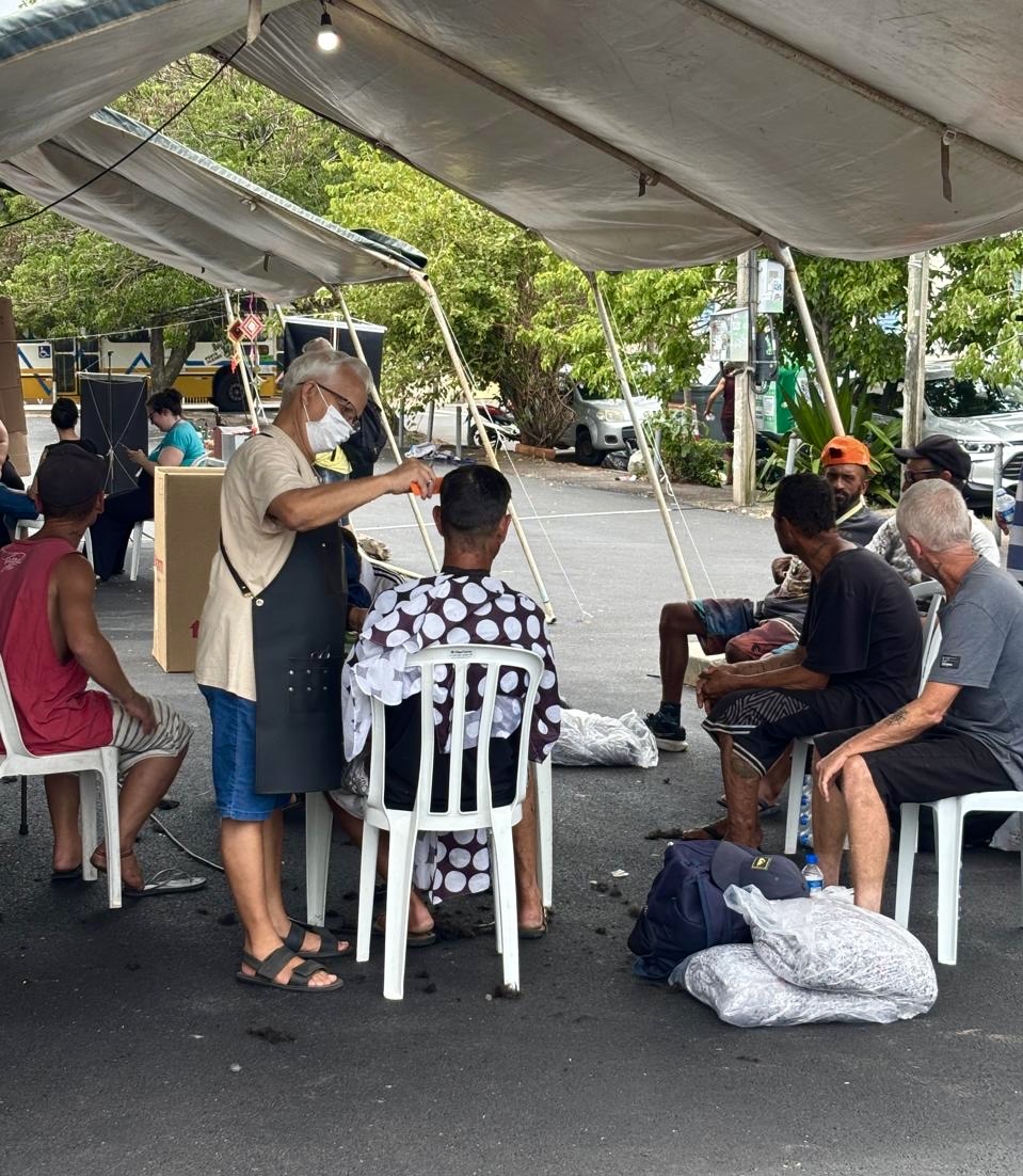 Em uma tenda, um profissional realiza corte de cabelo em uma pessoa sentada em cadeira branca, enquanto outras pessoas aguardam atendimento ao redor. Todos estão em ambiente aberto.