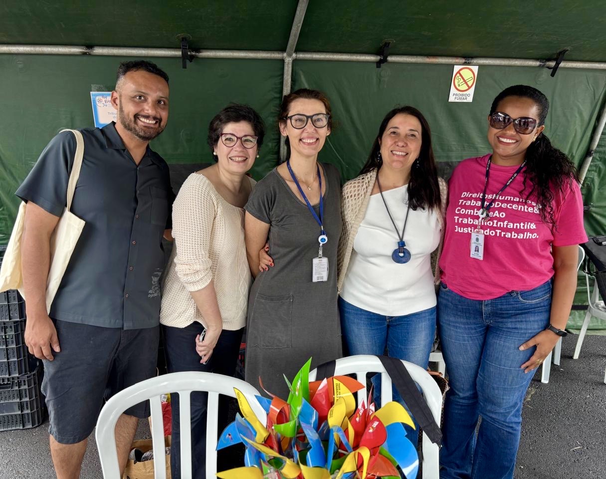Grupo de cinco pessoas posa para foto dentro de uma tenda do evento. Todos da Justiça do Trabalho, sorrindo, em frente a uma mesa com materiais coloridos.
