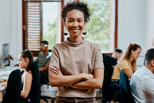 Foto ilustrativa de uma menina de pé com os braços cruzados sorrindo. ao fundo aparecem diversas pessoas sentadas em estações de trabalho, em um ambiente que lembra um escritório.