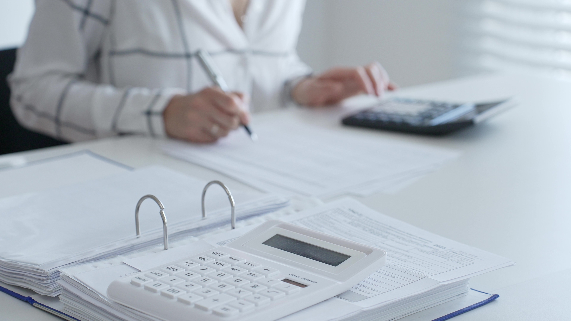 A imagem mostra uma pessoa de fundo segurando lápis, com uma calculadora em frente e mais na frente ainda uma agenda e outra calculadora. A imagem da pessoa está desfocada e sem aparecer o rosto.