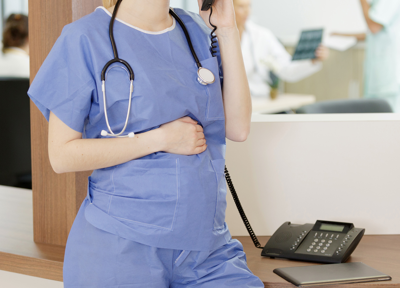 A foto mostra uma profissional de saúde grávida, com uniforme e estetoscópio no pescoço. Ela coloca uma das mãos na barriga.