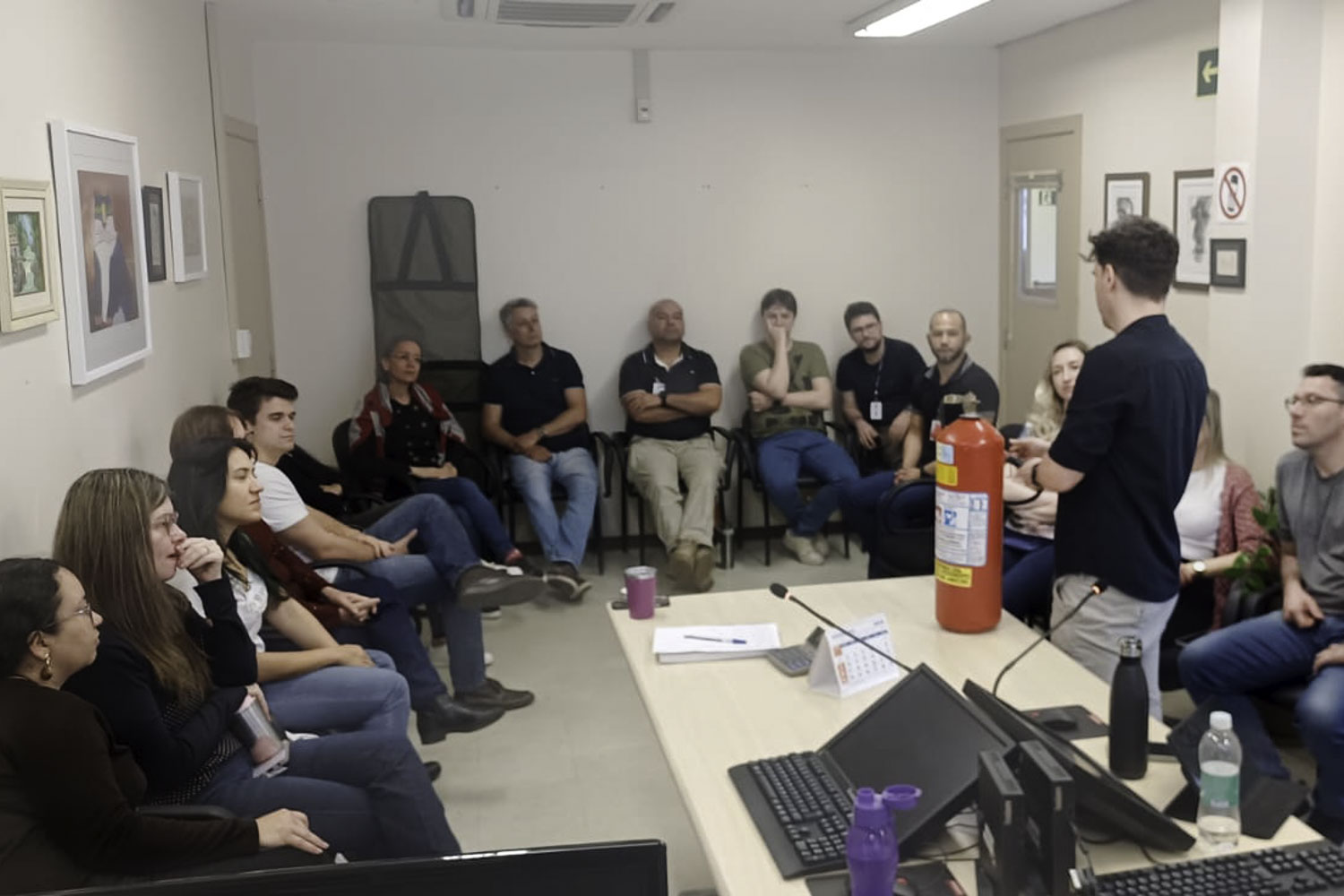 homem de pé fala para um círculo de pessoas sentadas. há uma mesa com um extintor sobre ela. 