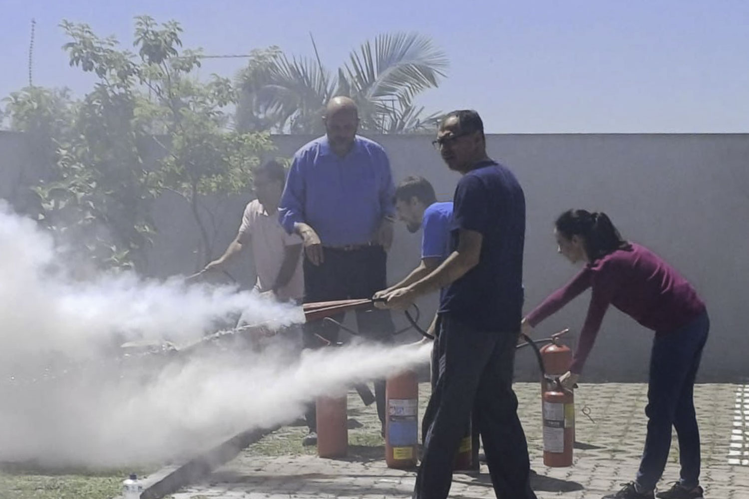 cinco pessoas aparecem na foto, segurando três extintores que expelem fumaça branca. Eles estão no pátio, ao ar livre. 