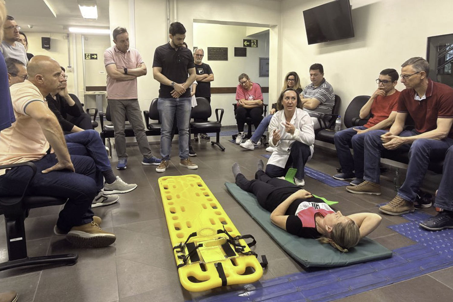 uma mulher está deitada no chão e outra está de joelhos, usando jaleco, dando explicações para um grupo de pessoas que estão sentadas em círculo