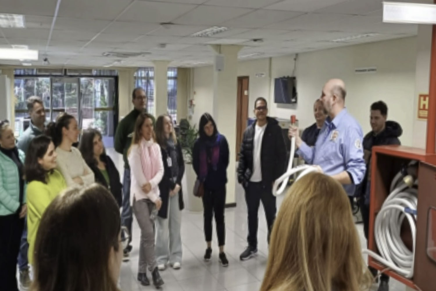 Homem de pé segura uma mangueira de incêndio e fala para um grupo que está em círculo ouvindo
