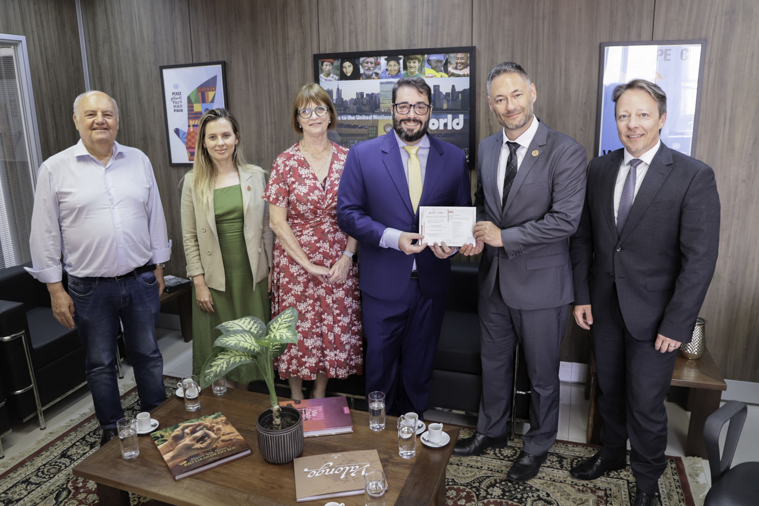 seis pessoas posam para a foto de pé e sorrindo. O presidente do TRT e o presidente eleito da Ajuris aparecem segurando o convite da posse.