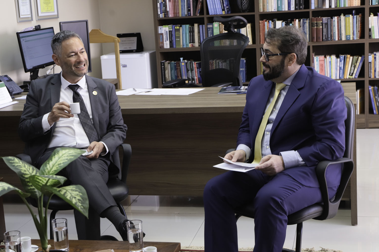 Dois homens aparecem sentados, conversando e sorrindo. eles seguram xícaras de café. 