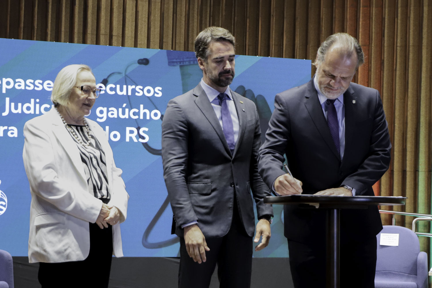 Três pessoas estão de pé no palco. Na direita, Alberto Delgado assina o termo de repasse. 