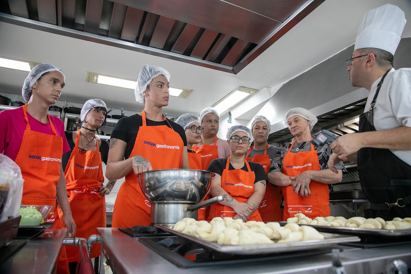 Foto de alunos(a) do curso ouvindo as instruções do professor, em uma cozinha industrial