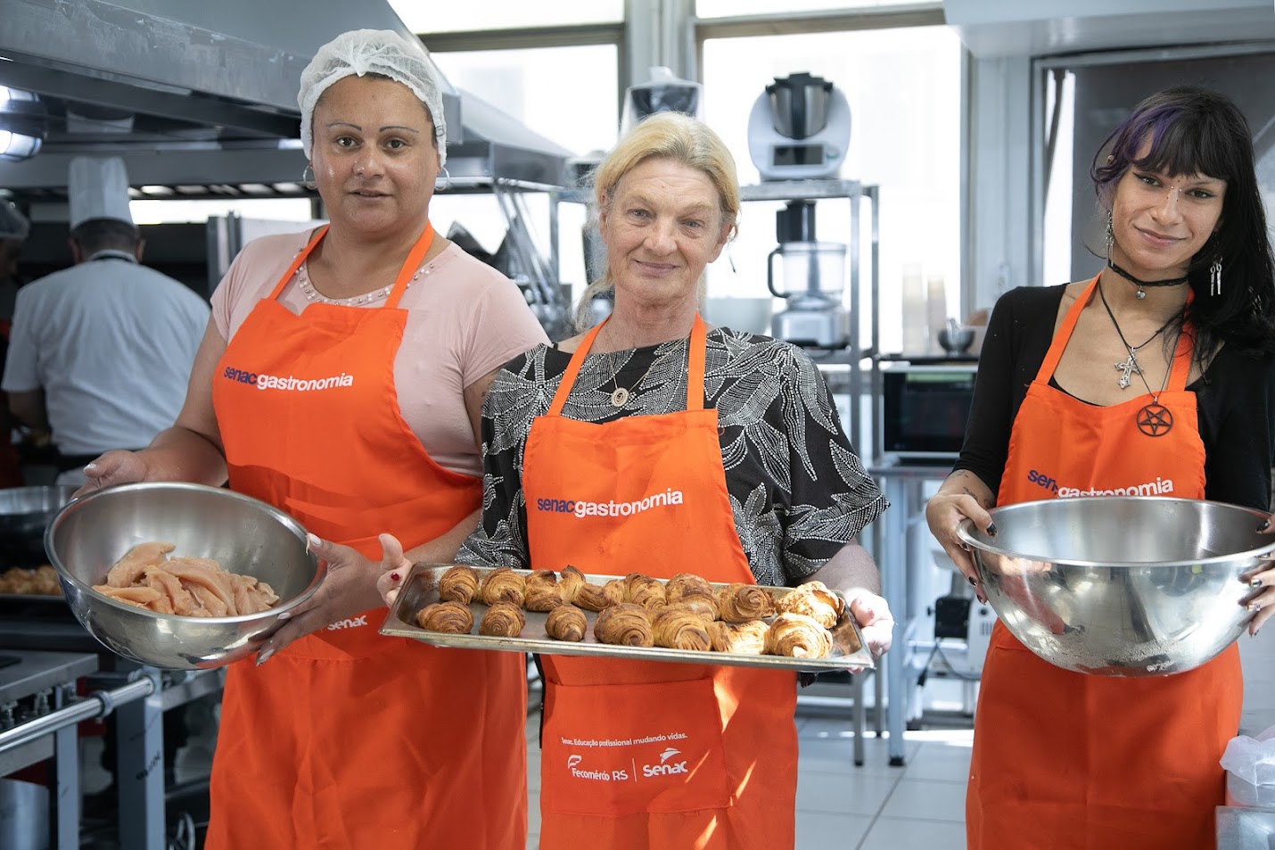 Foto posada de três alunas do curso, segurando recipientes com alimentos em preparação.