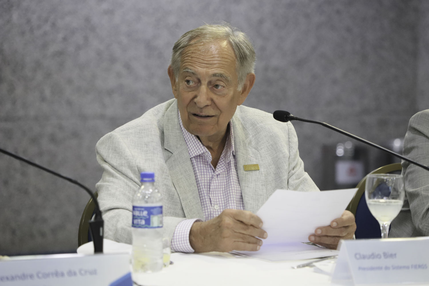 Cláudio Bier é branco, calvo. Usa camisa xadrez e blazer claro. Fala e olha para a esquerda.