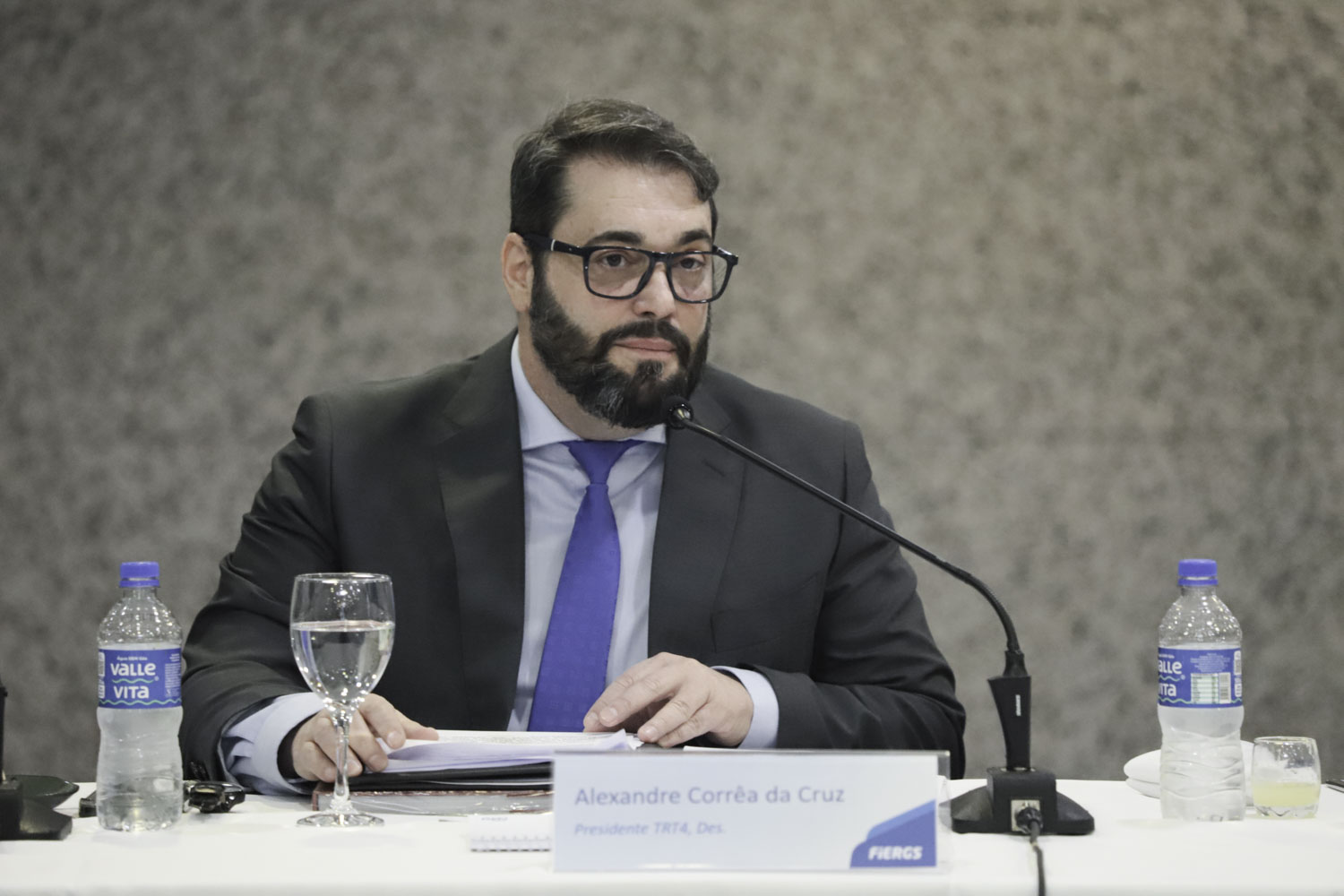 Alexandre é branco, tem cabelo e barba preta e usa óculos de aro grosso. veste terno reto e camisa e gravata azuis.