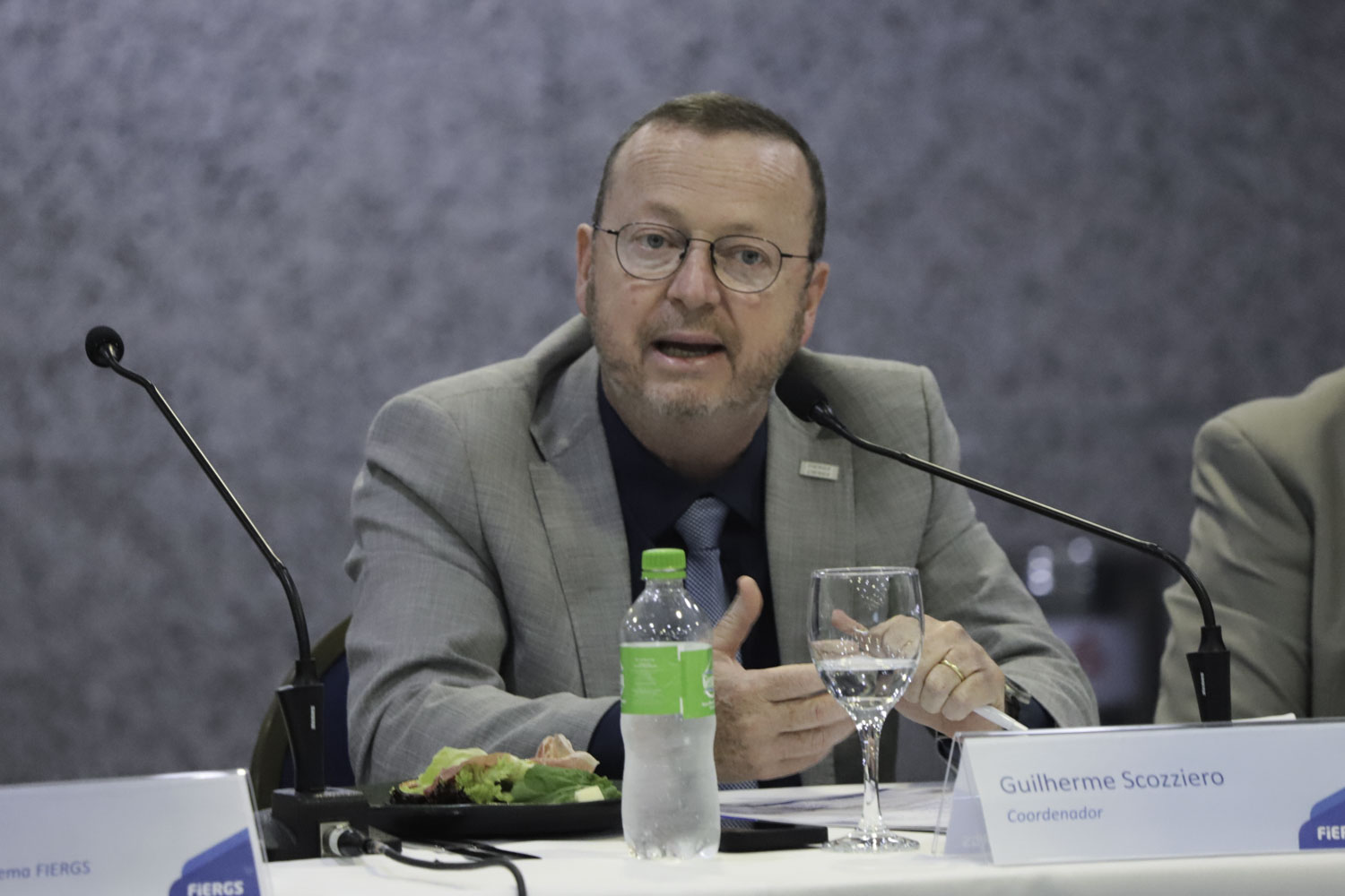 Scozziero é branco e ruivo. Usa blazer cinza, camisa preta e gravata cinza. Fala e gesticula olhando para a direita. 