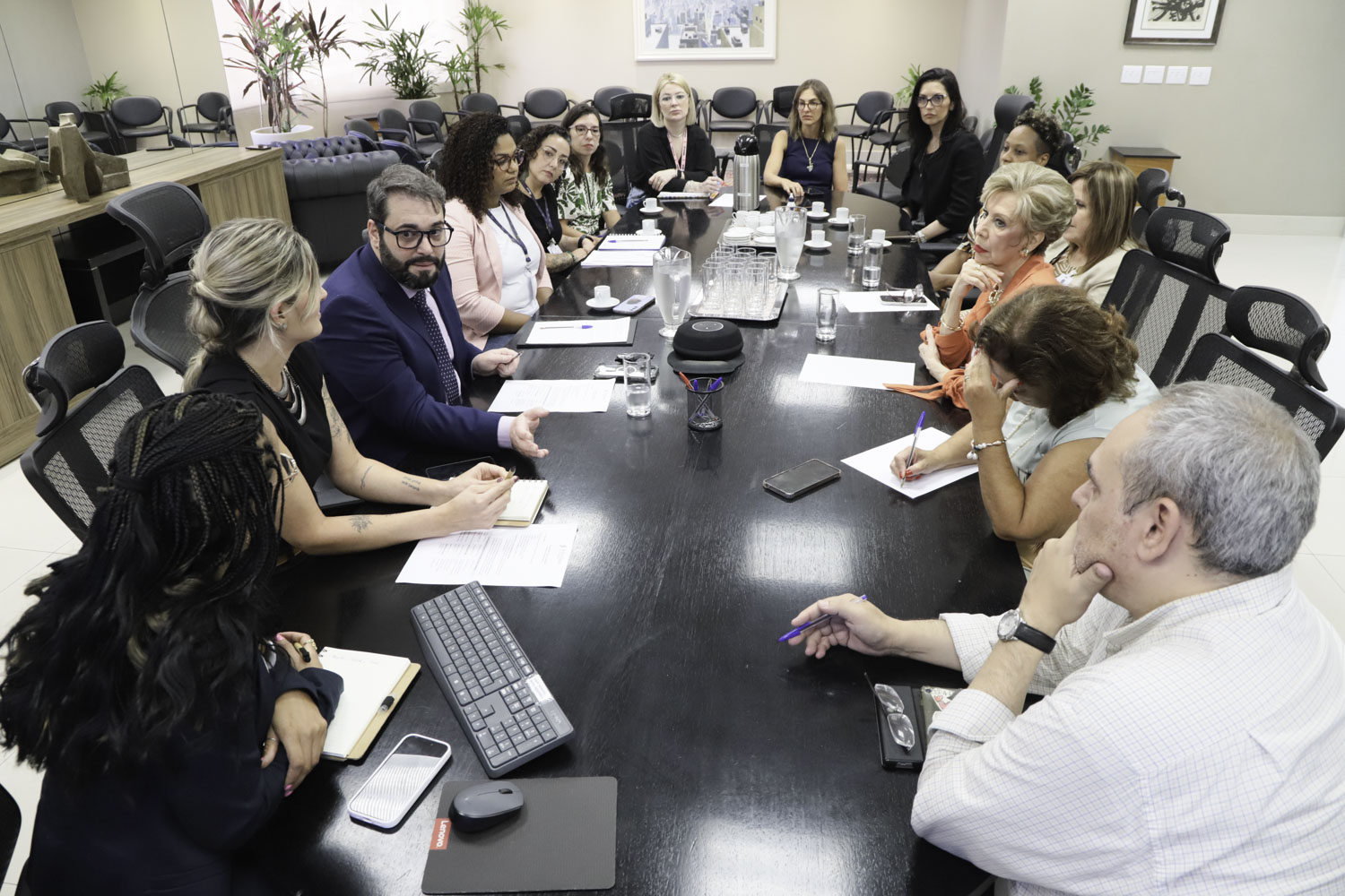 visão da reunião pelo ângulo oposto, com as pessoas ao redor da mesa e o presidente falando na esquerda da imagem. 