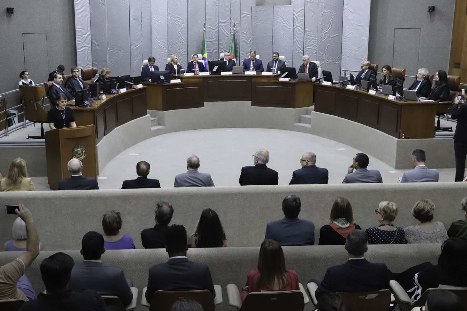 mesa oficial ao fundo com público de costas em primeiro plano