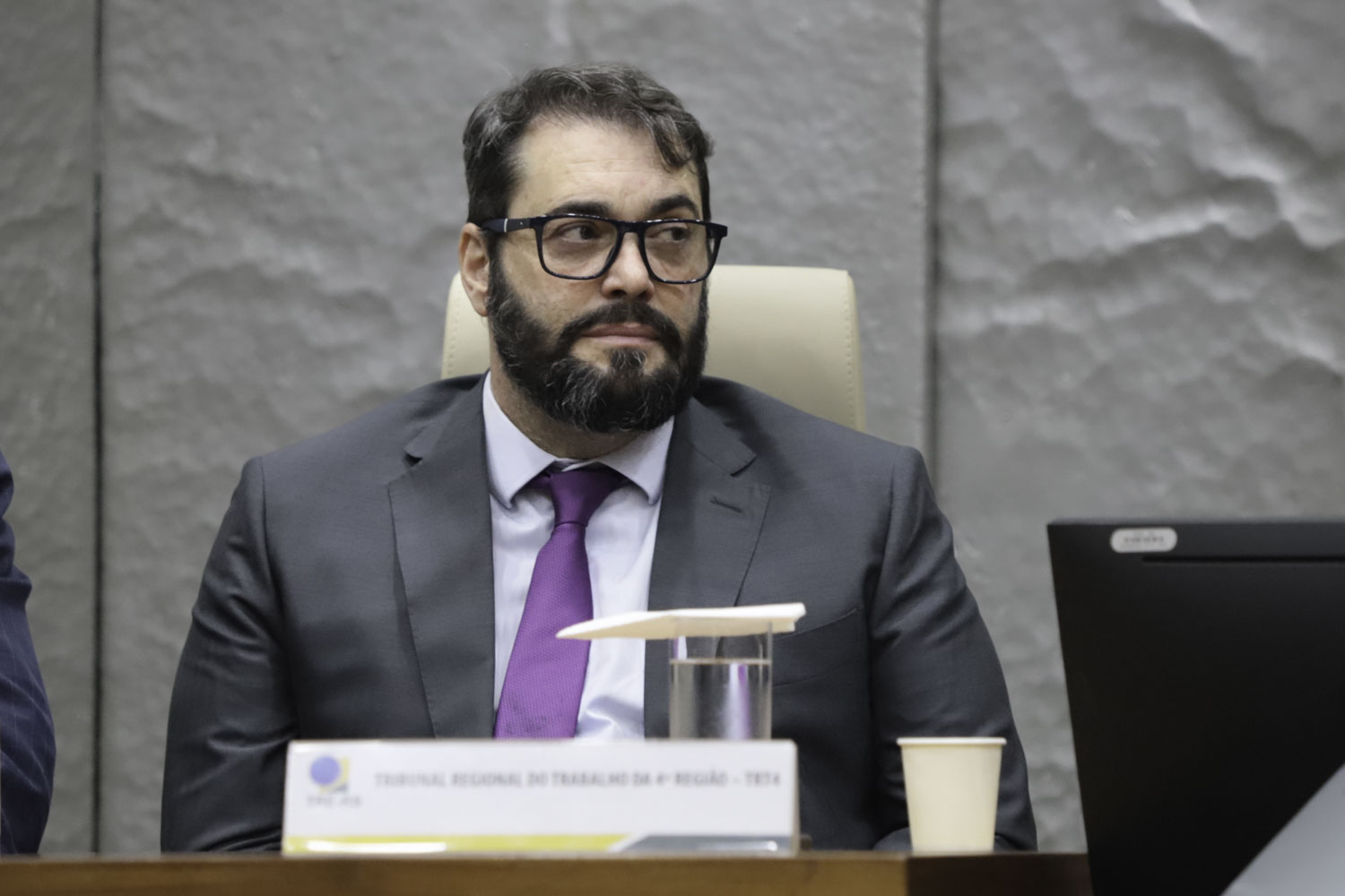 Alexandre é branco, de cabelo e barba pretos. Usa terno cinza, com camisa branca e gravata lilás, e óculos de aro preto. Está olhando para a direita. 